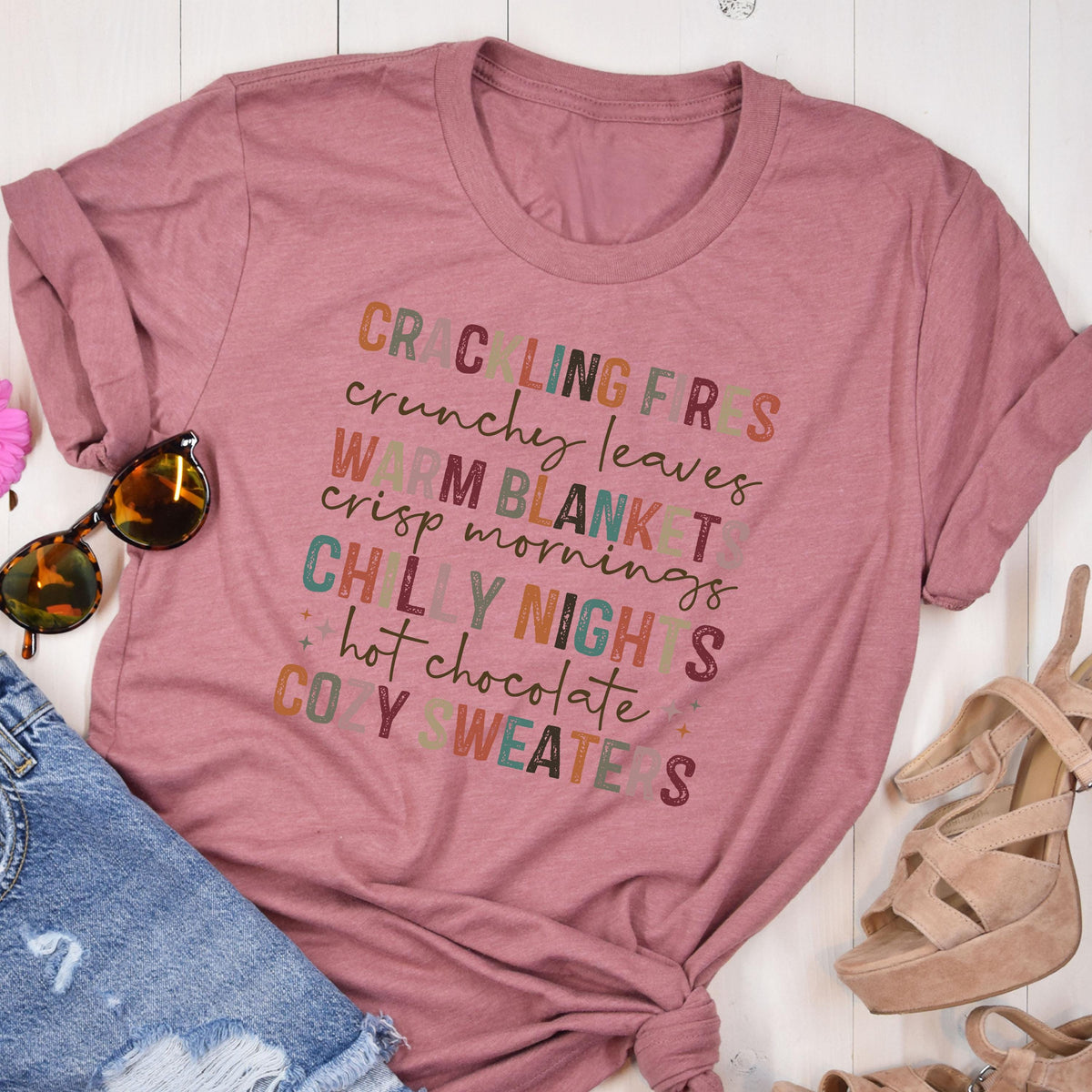 May include: A dusty rose-colored t-shirt with rolled-up sleeves. The shirt features colorful text that reads "Crackling Fires, Crunchy Leaves, Warm Blanket, Crisp Mornings, Chilly Nights, Hot Chocolate, Cozy Sweaters."