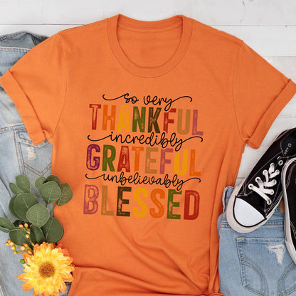 May include: An orange short-sleeved t-shirt featuring the text "so very THANKFUL incredibly GRATEFUL unbelievably BLESSED" in a mix of black cursive and colorful, distressed block lettering. The shirt is displayed with blue denim and a yellow flower.