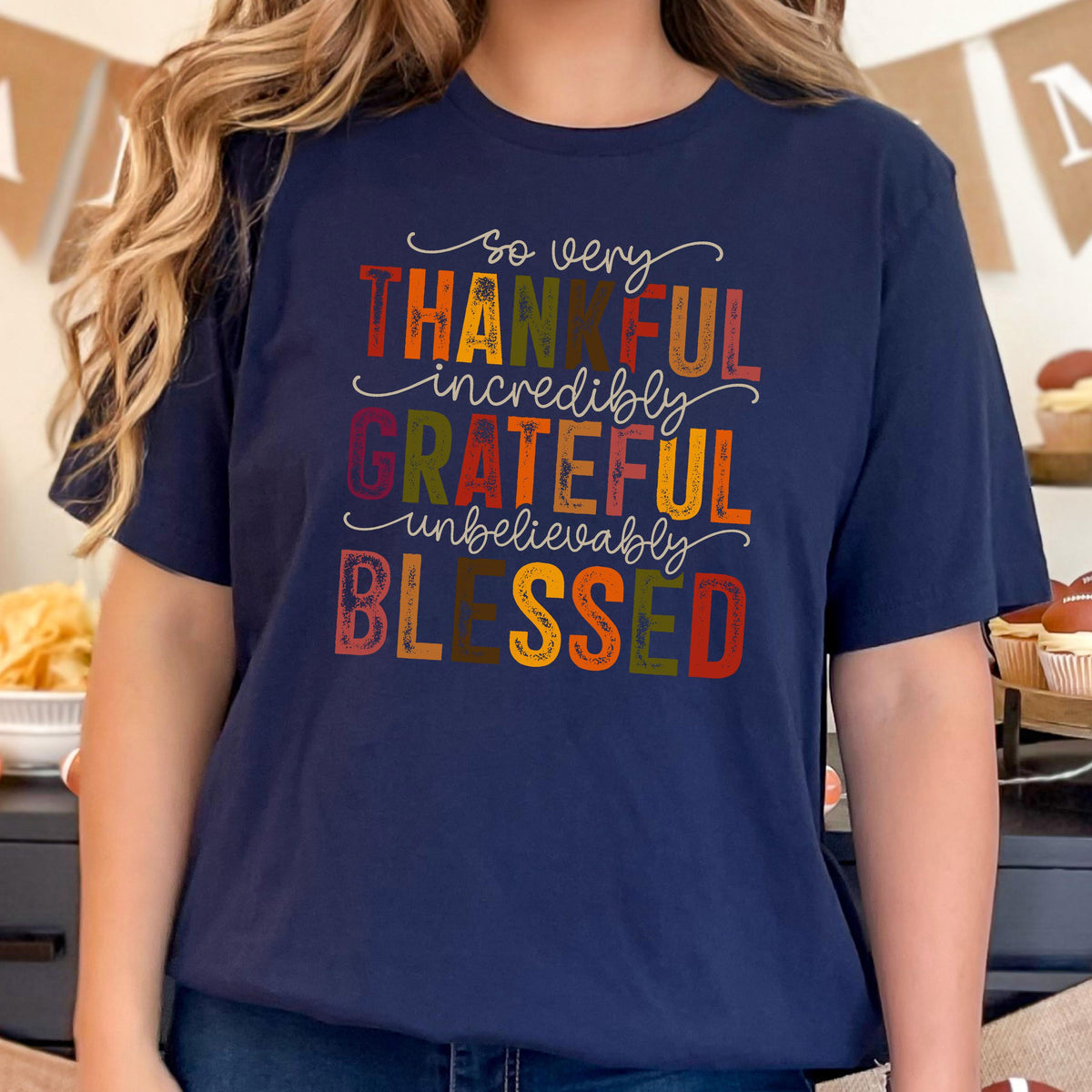 May include: A navy blue short-sleeve t-shirt featuring the text "so very THANKFUL incredibly GRATEFUL unbelievably BLESSED" in a multi-colored font with shades of orange, yellow, green, and red, arranged in a stacked, stylized design.