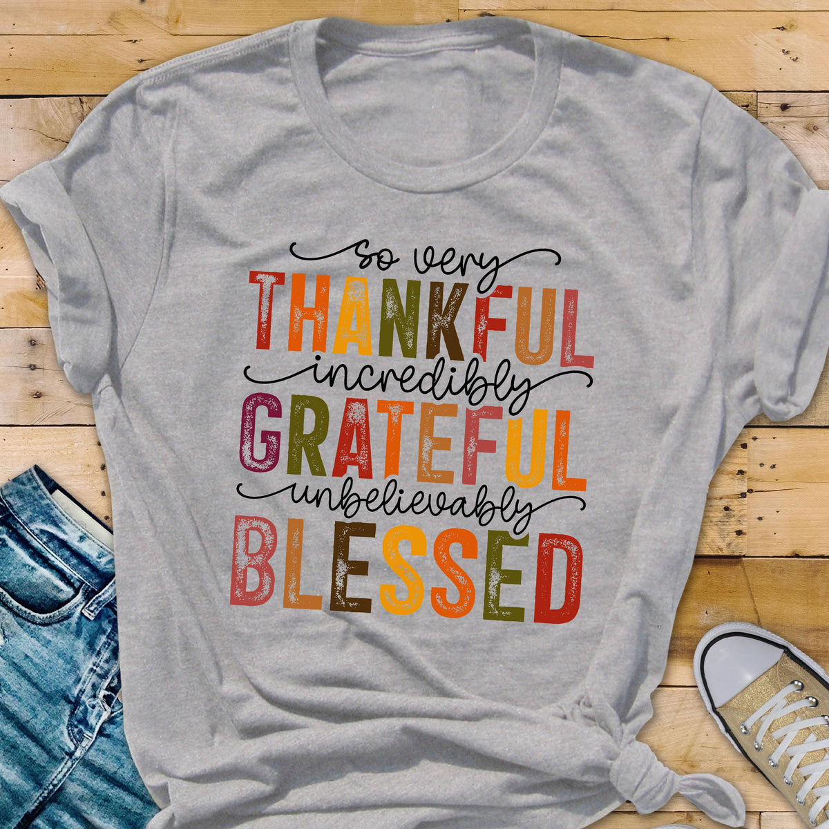 May include: A light gray short-sleeved t-shirt featuring the text so very THANKFUL incredibly GRATEFUL unbelievably BLESSED in a multi-colored, distressed font with shades of red, orange, yellow, green, and brown on the front of the garment.