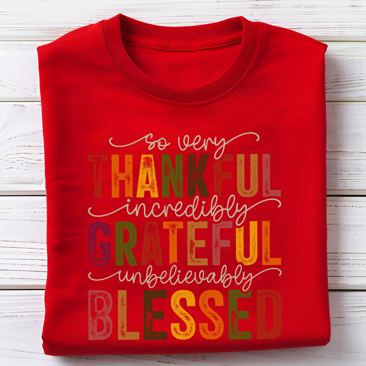 May include: A bright red crew neck t-shirt featuring a multi-colored graphic print with the text so very THANKFUL incredibly GRATEFUL unbelievably BLESSED. The lettering uses a mix of script and block fonts in shades of orange, yellow, green, and brown.