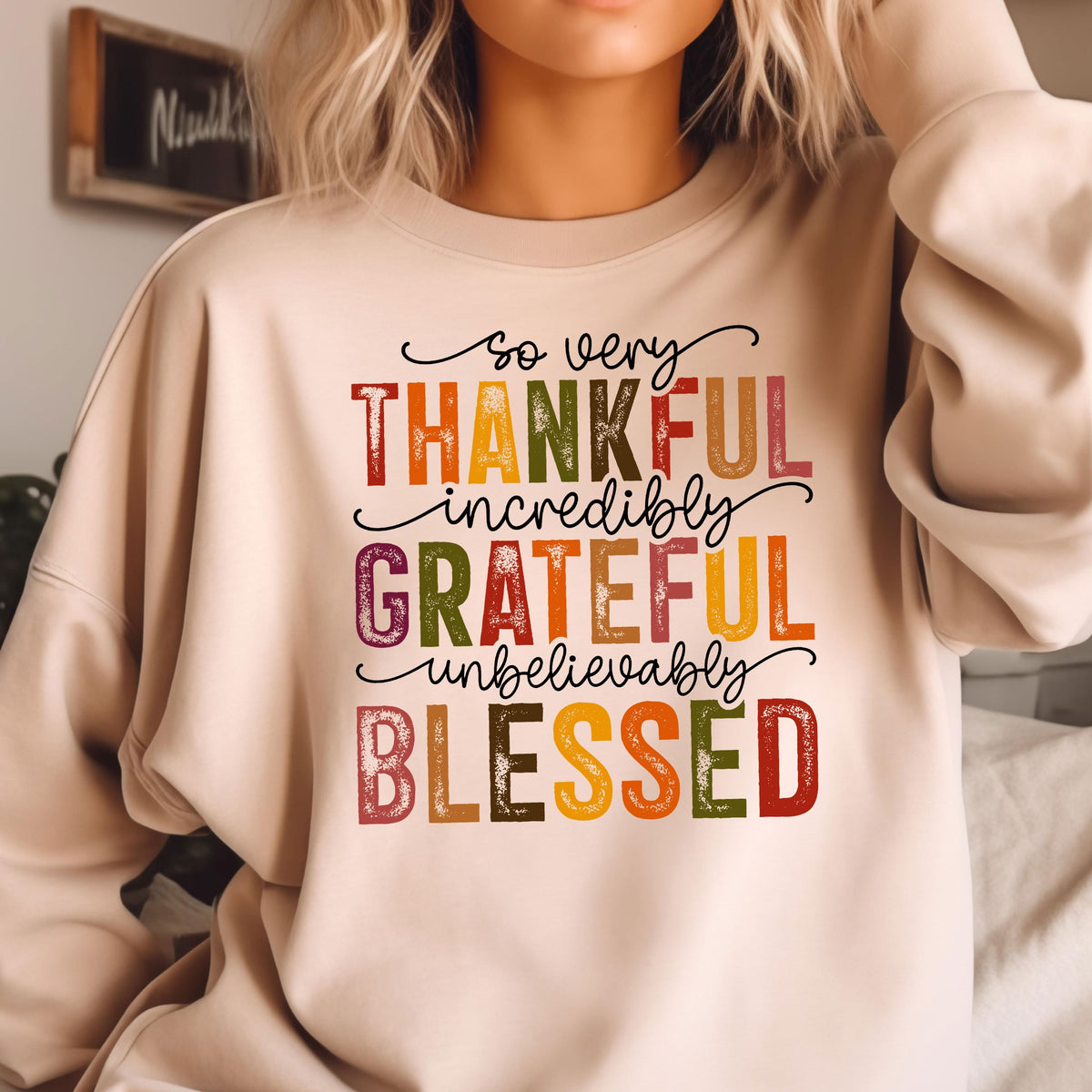 May include: A beige crewneck sweatshirt featuring the text so very THANKFUL incredibly GRATEFUL unbelievably BLESSED in a mix of cursive and block lettering. The words are printed in various autumn-inspired colors including red, orange, yellow, and green.