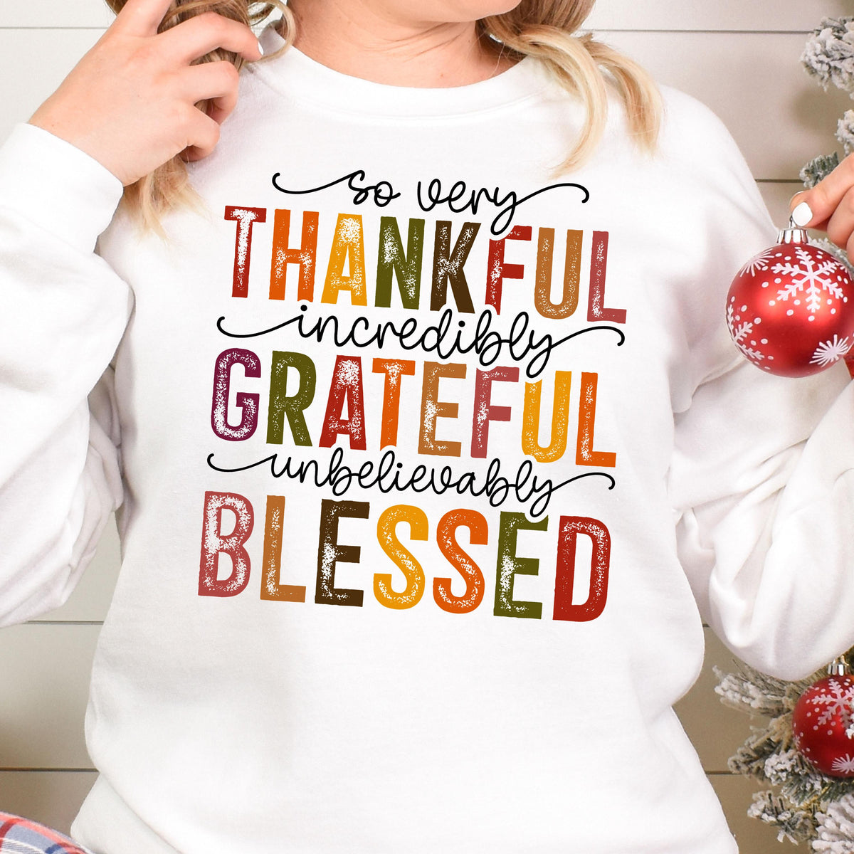 May include: A white long-sleeve sweatshirt featuring the text "so very THANKFUL incredibly GRATEFUL unbelievably BLESSED" in a multi-colored, distressed font. The words are arranged in a stacked design with a mix of cursive and block lettering.