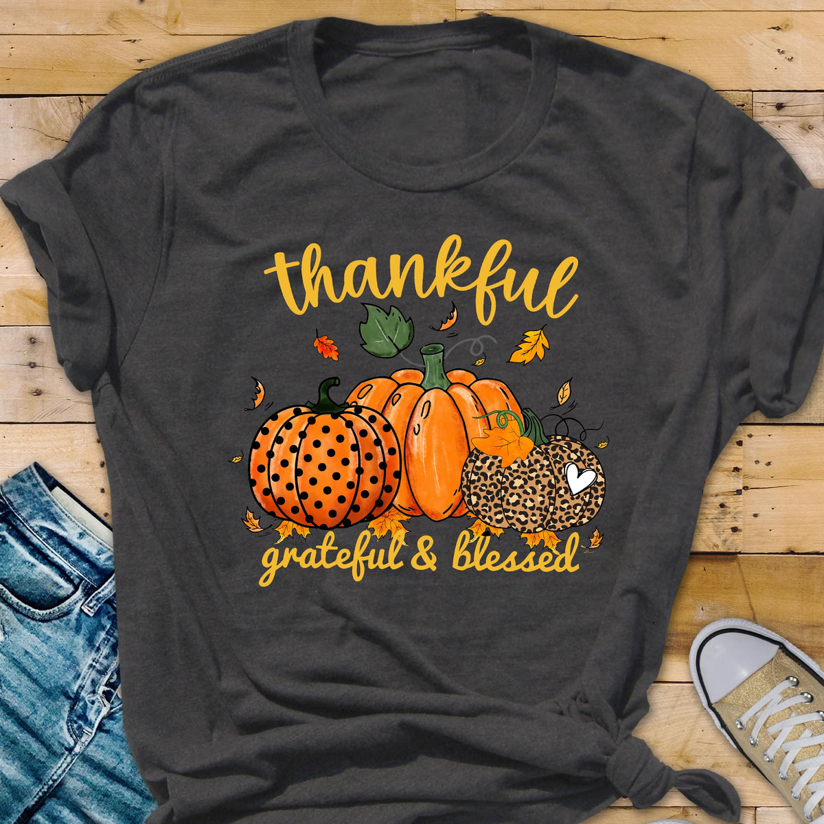 May include: Dark gray t-shirt with a fall-themed graphic. The design features three pumpkins in orange and leopard print, with the words "thankful" and "grateful & blessed" in yellow script. Autumn leaves are scattered around the pumpkins.