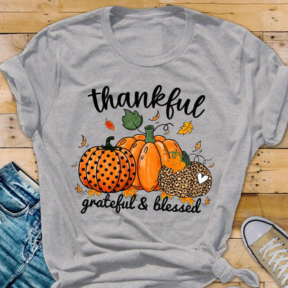 May include: A heather gray t-shirt featuring a fall-themed graphic. The design includes three pumpkins, fall leaves, and the words "thankful, grateful & blessed" in black script. The pumpkins are orange and one has a leopard print.