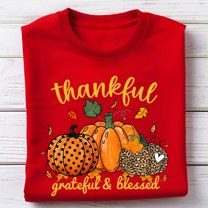 May include: A red sweatshirt featuring a fall-themed design. The design includes three pumpkins, fall leaves, and the words "thankful, grateful & blessed" in yellow script. One pumpkin has an orange polka dot pattern, another is solid orange, and the third has a leopard print.