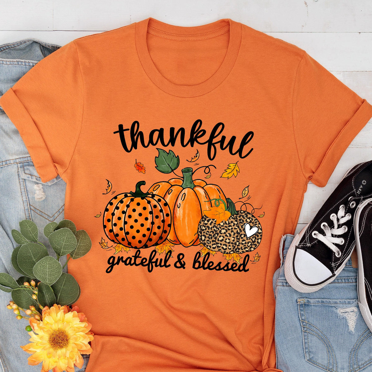 May include: Orange t-shirt with a fall-themed graphic. The design features three pumpkins, one with polka dots, one solid orange, and one with a leopard print pattern. The words "thankful, grateful & blessed" are written in black script.