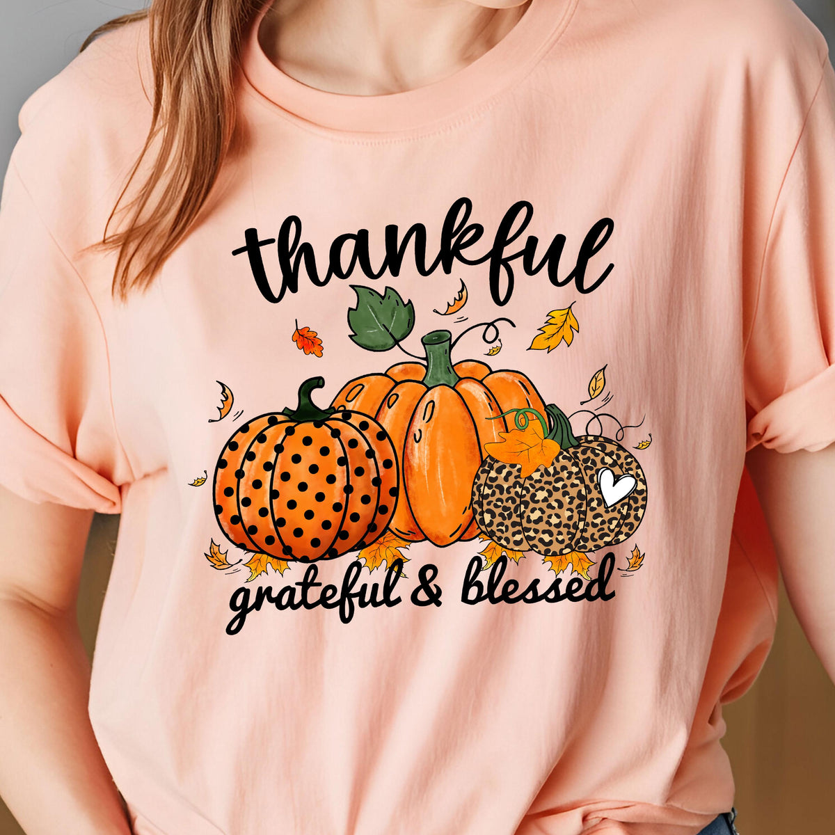 May include: Peach-colored t-shirt featuring a fall-themed graphic. The design includes three pumpkins in orange and leopard print, with the words "thankful, grateful & blessed" in black script. Autumn leaves are scattered around the pumpkins.