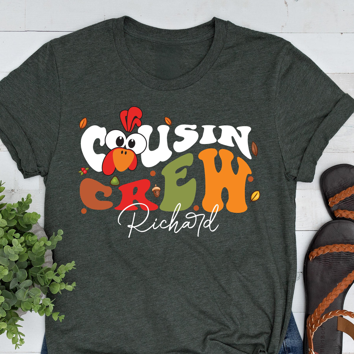May include: A dark charcoal gray t-shirt featuring a graphic with the text "COUSIN CREW" in large, colorful, retro-style bubble letters. A cartoon turkey head is integrated into the text, and the name "Richard" is written below in white script.