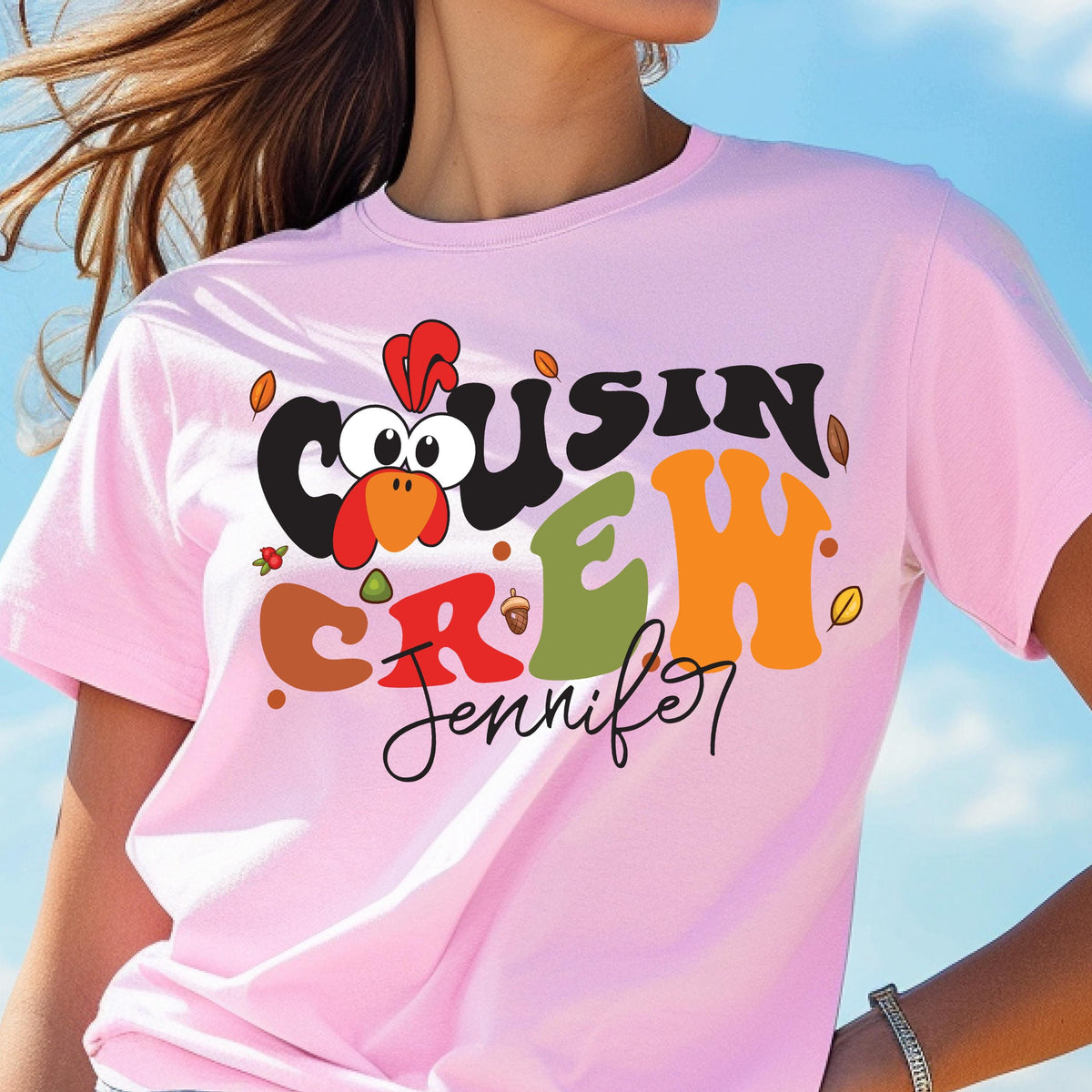 May include: A light pink short-sleeve t-shirt featuring a graphic with the text "COUSIN CREW" in black, olive green, and orange, a cartoon turkey head, and the name "Jennifer" in black cursive script, accented with small colorful autumn leaves.