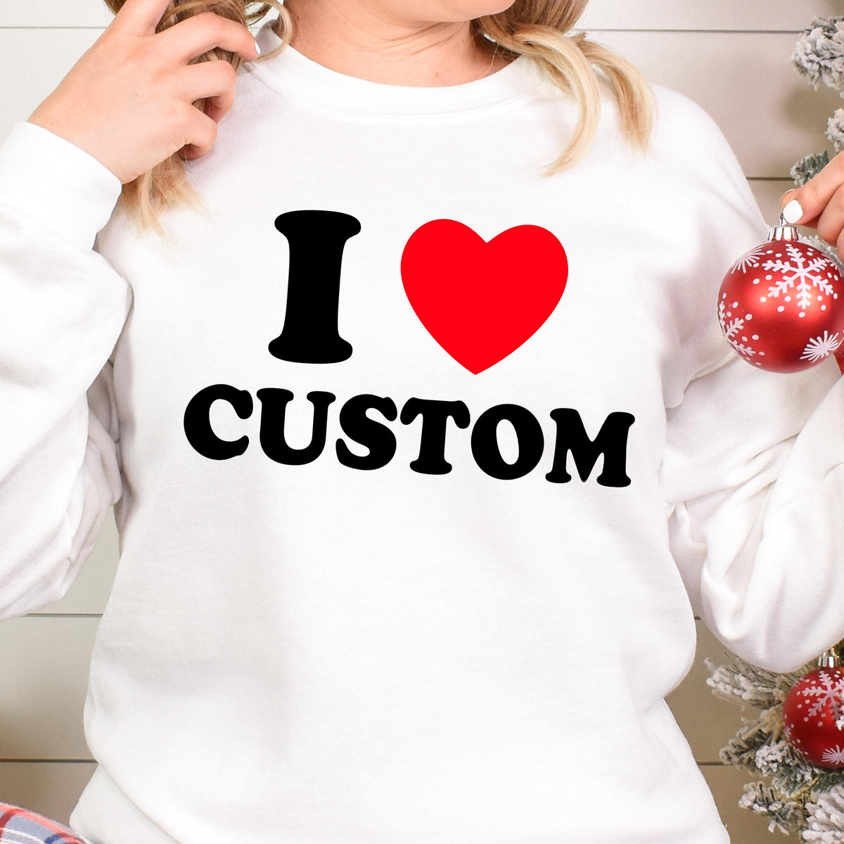 May include: White sweatshirt with the text "I ❤ CUSTOM" in black and a red heart. The sweatshirt is a long-sleeved crew neck. A Christmas ornament is held in the right hand.