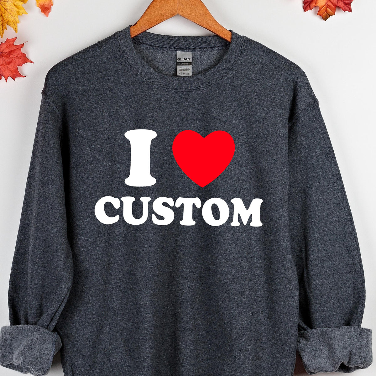 May include: Dark gray sweatshirt with the text "I ♥ CUSTOM" in white and a red heart. The sweatshirt is displayed on a wooden hanger. The design is centered on the front of the garment.