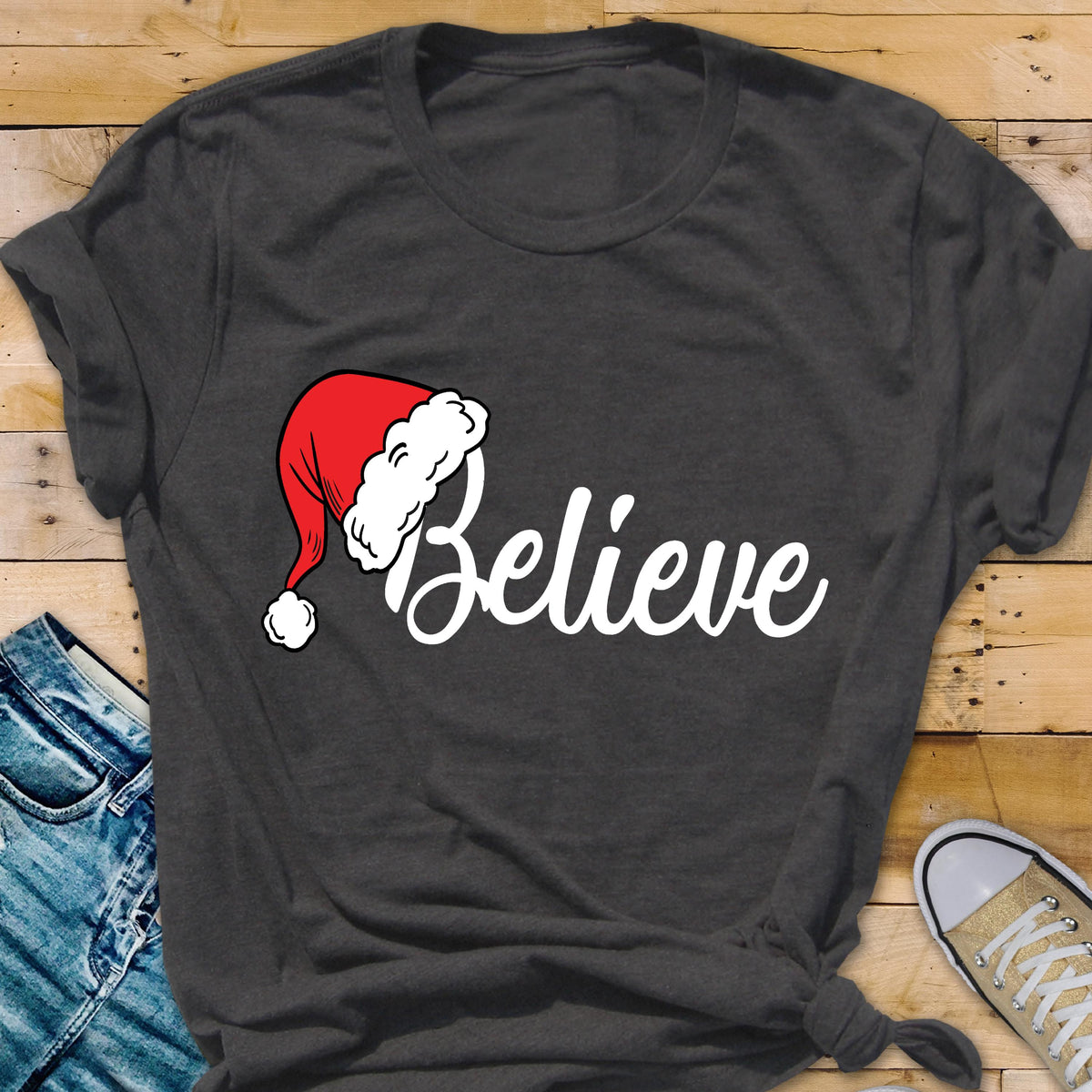 May include: A dark charcoal gray short-sleeved t-shirt featuring the word Believe in white cursive script. The letter B is replaced by a red and white Santa Claus hat with a white pom-pom at the tip, centered on the front of the garment.