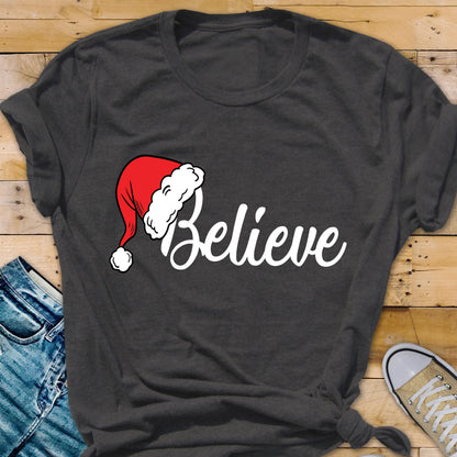 May include: A dark charcoal gray short-sleeved t-shirt featuring the word Believe in white cursive script. The letter B is replaced by a red and white Santa Claus hat with a white pom-pom at the tip, centered on the front of the garment.