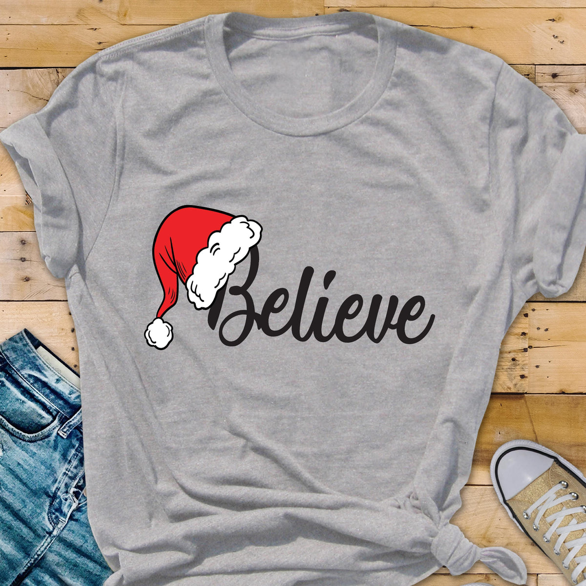 May include: A light gray short-sleeved t-shirt featuring the word Believe in black cursive script. A red and white Santa hat is positioned over the letter B. The shirt has a crew neckline and is made of a soft, heathered fabric material.