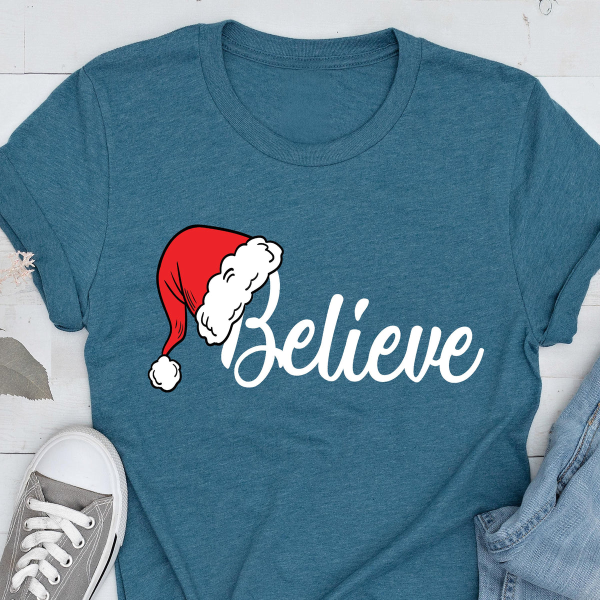 May include: A teal short-sleeved t-shirt featuring the word Believe in white script lettering. The letter B is replaced by a red Santa hat with a white pom-pom and a white fur trim. The shirt has a crew neckline and a heathered fabric texture.