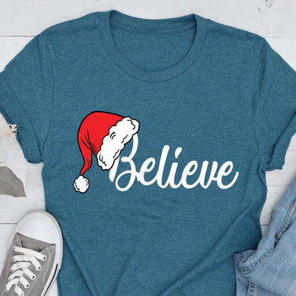 May include: A teal short-sleeved t-shirt featuring the word Believe in white script lettering. The letter B is replaced by a red Santa hat with a white pom-pom and a white fur trim. The shirt has a crew neckline and a heathered fabric texture.