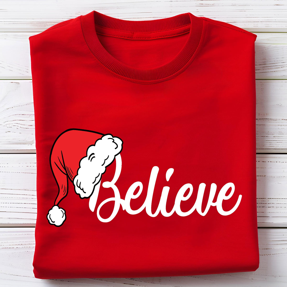 May include: A bright red crew neck t-shirt featuring a white graphic of a Santa hat positioned above the word Believe written in a white cursive script font. The shirt is folded neatly, showcasing the front design clearly on the fabric surface.