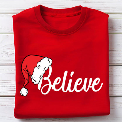 May include: A bright red crew neck t-shirt featuring a white graphic of a Santa hat positioned above the word Believe written in a white cursive script font. The shirt is folded neatly, showcasing the front design clearly on the fabric surface.