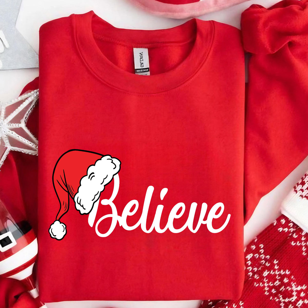 May include: A bright red crewneck sweatshirt featuring a white graphic of a Santa hat and the word Believe in a white cursive script font. The garment has a ribbed collar and a standard long-sleeve design with a visible interior brand label.