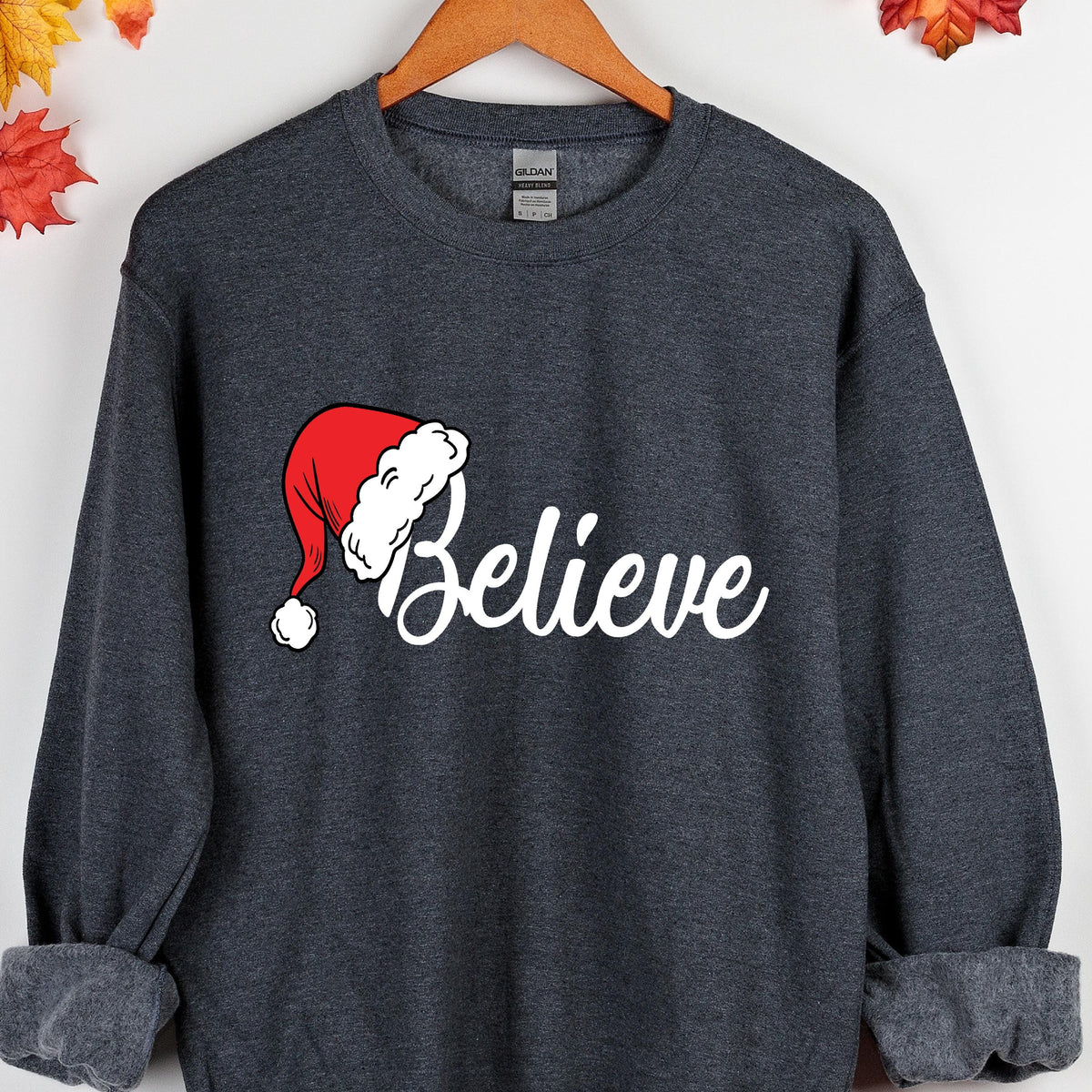 May include: A dark charcoal gray crewneck sweatshirt featuring a red and white Santa hat graphic positioned above the white cursive text "Believe" printed on the center chest. The garment has long sleeves with ribbed cuffs and a ribbed neckline.