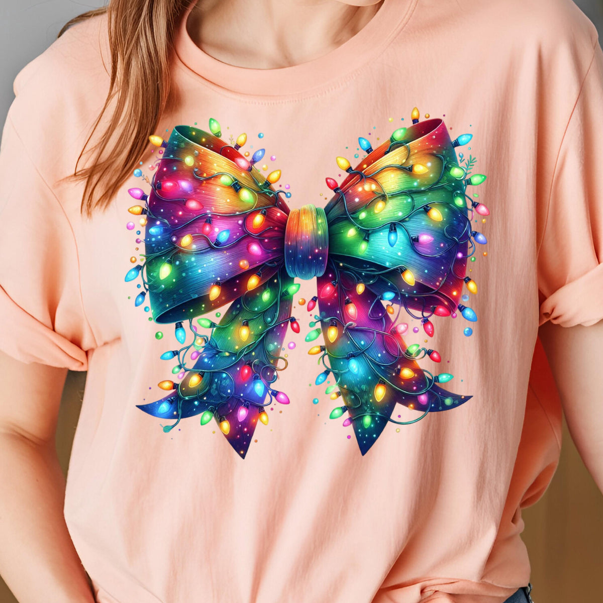 May include: A light pink t-shirt featuring a large, colorful bow design. The bow is decorated with a string of Christmas lights in various colors, including red, green, blue, and yellow. The bow is the focal point of the shirt.