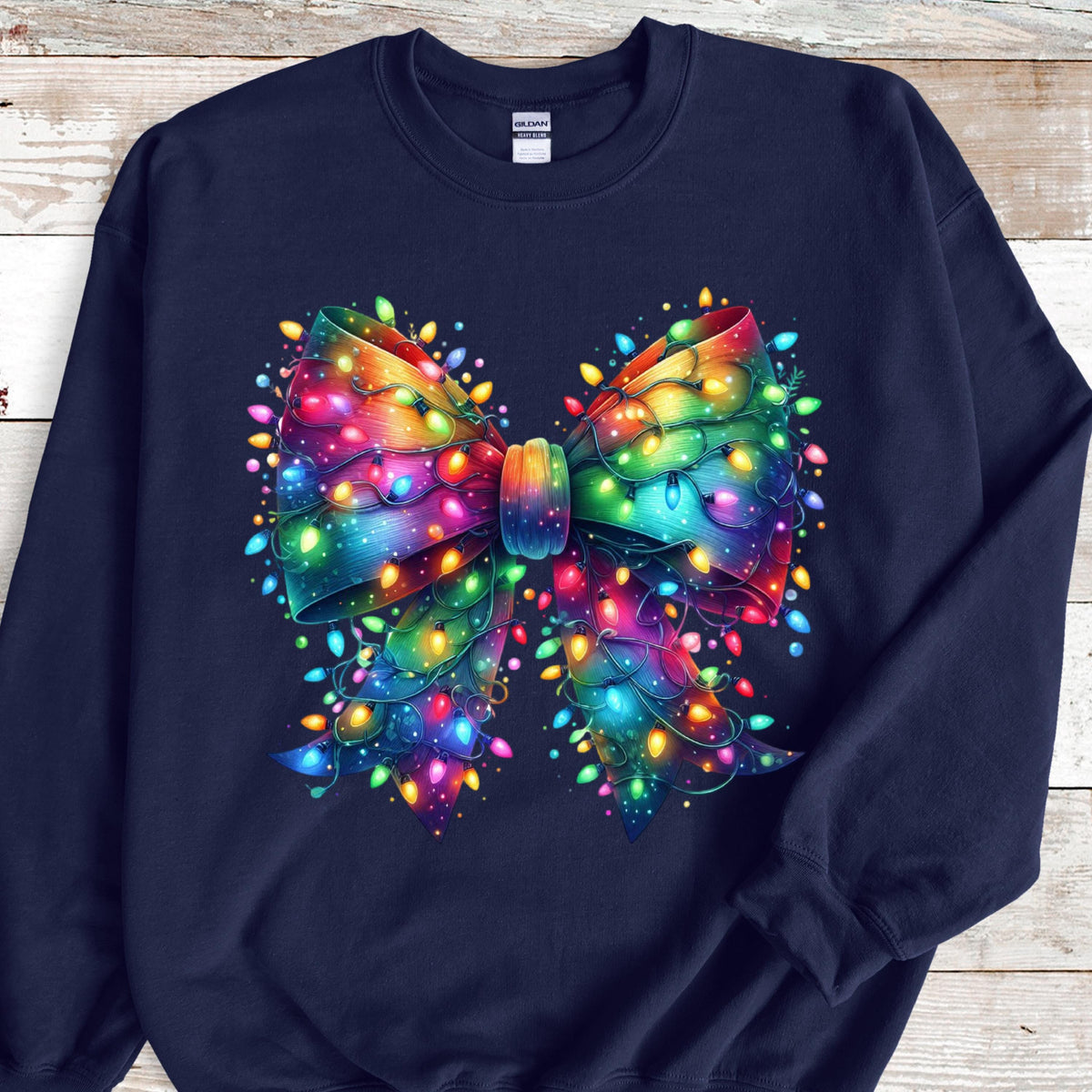 May include: A navy blue crewneck sweatshirt featuring a large, central graphic of a decorative bow. The bow is rendered in a vibrant, multi-colored gradient and is intricately wrapped with glowing, multi-colored string Christmas lights.