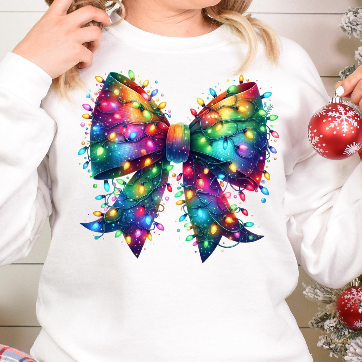 May include: A white long-sleeve sweatshirt featuring a large, central graphic of a bow made from colorful, glowing Christmas string lights in shades of blue, green, yellow, red, and purple, with a red ornament visible on the right side.