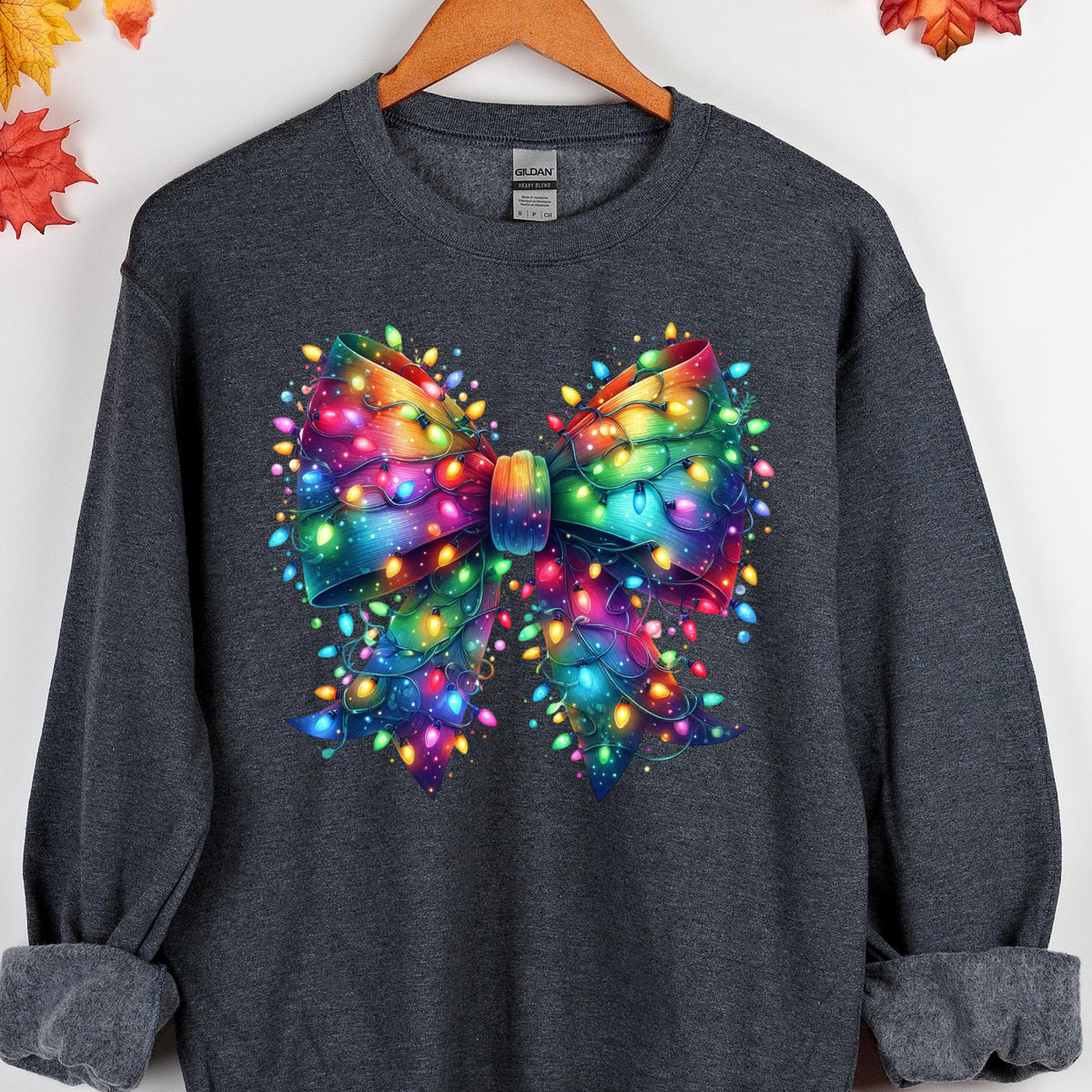 May include: A dark charcoal gray long-sleeve sweatshirt featuring a large, central graphic of a bow made entirely of glowing, multi-colored Christmas string lights. The lights appear in vibrant shades of red, blue, green, yellow, and purple.