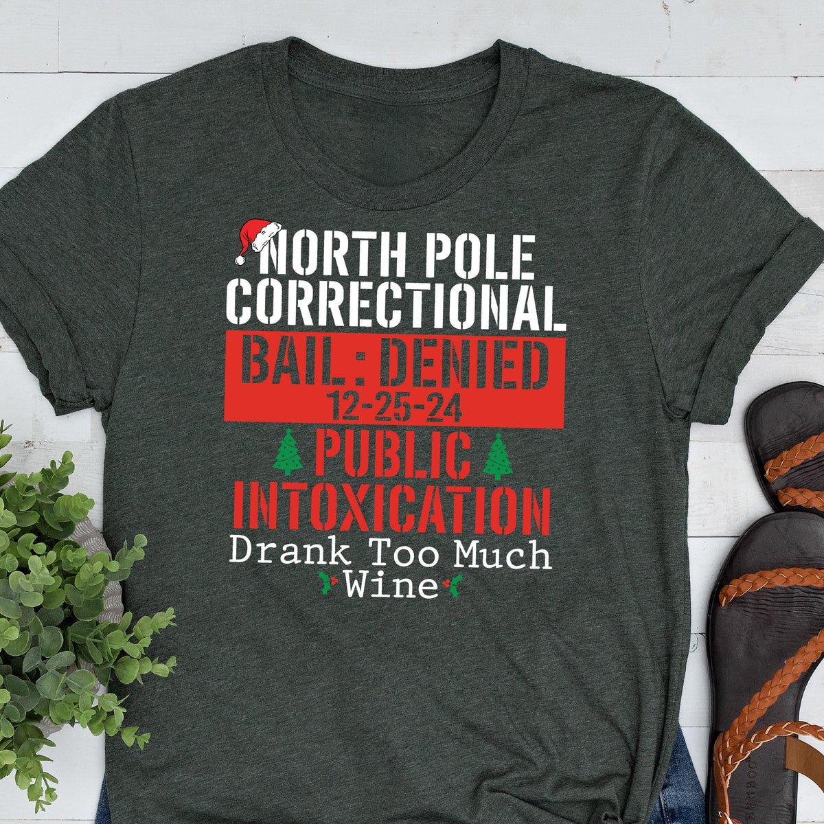 May include: A dark heather gray t-shirt featuring white and red text that reads NORTH POLE CORRECTIONAL BAIL: DENIED 12-25-24 PUBLIC INTOXICATION Drank Too Much Wine, accented with a small red Santa hat icon and two green Christmas tree graphics.