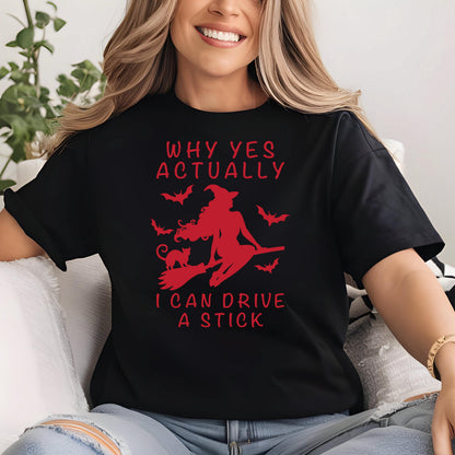 May include: Black t-shirt with a red graphic of a witch on a broomstick, a black cat, and bats. The text "WHY YES ACTUALLY I CAN DRIVE A STICK" is printed in red. The shirt is made of a soft material.