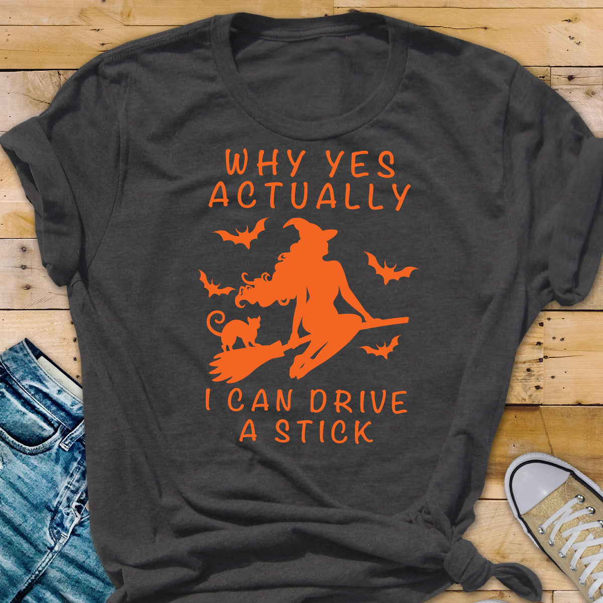 May include: Dark gray t-shirt with orange graphic of a witch on a broom, a cat, and bats. Text reads "WHY YES ACTUALLY I CAN DRIVE A STICK."