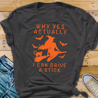 May include: Dark gray t-shirt with orange graphic of a witch on a broom, a cat, and bats. Text reads "WHY YES ACTUALLY I CAN DRIVE A STICK."