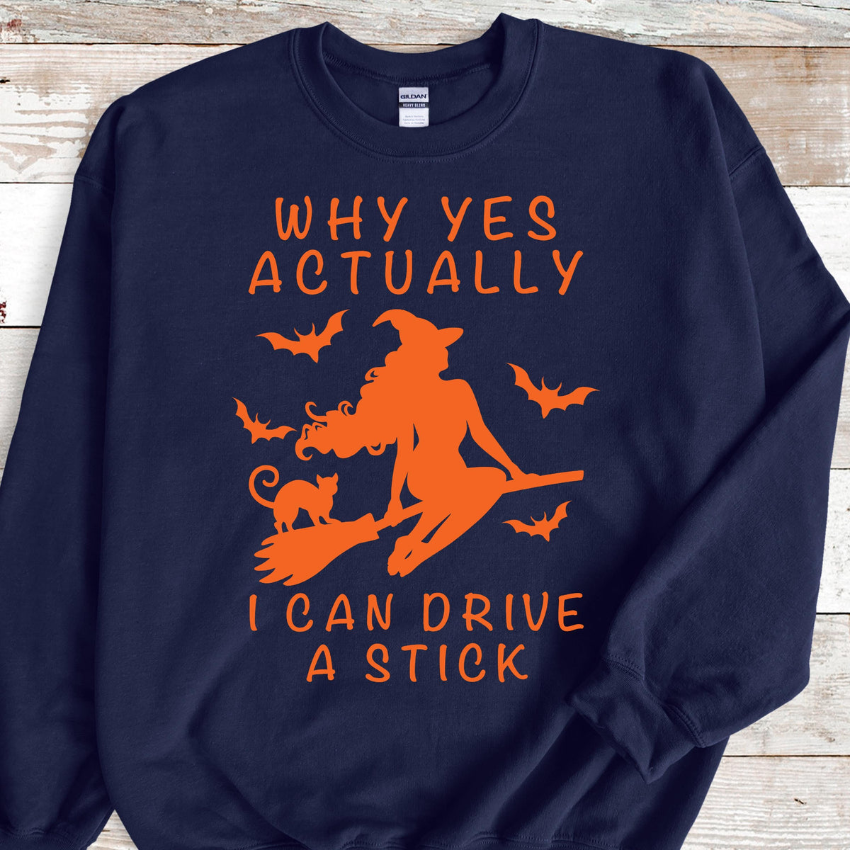 May include: A navy blue crewneck sweatshirt featuring an orange graphic of a witch riding a broomstick with a cat and bats. The text reads: WHY YES ACTUALLY I CAN DRIVE A STICK. The design is centered on the front of the long-sleeved pullover garment.