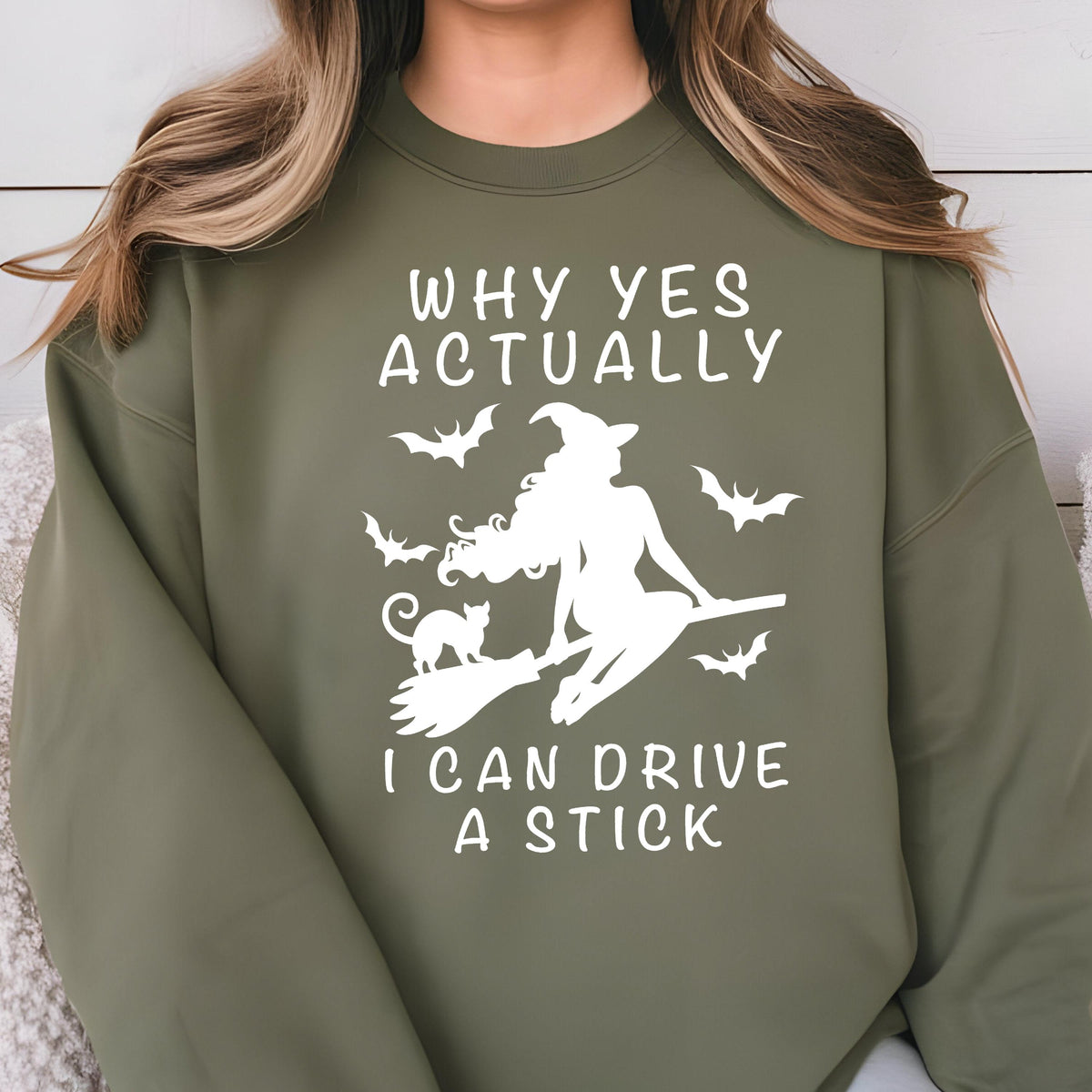 May include: An olive green crewneck sweatshirt featuring a white graphic print of a witch riding a broomstick with a cat and bats. The text reads, "WHY YES ACTUALLY I CAN DRIVE A STICK," positioned above and below the central witch illustration.