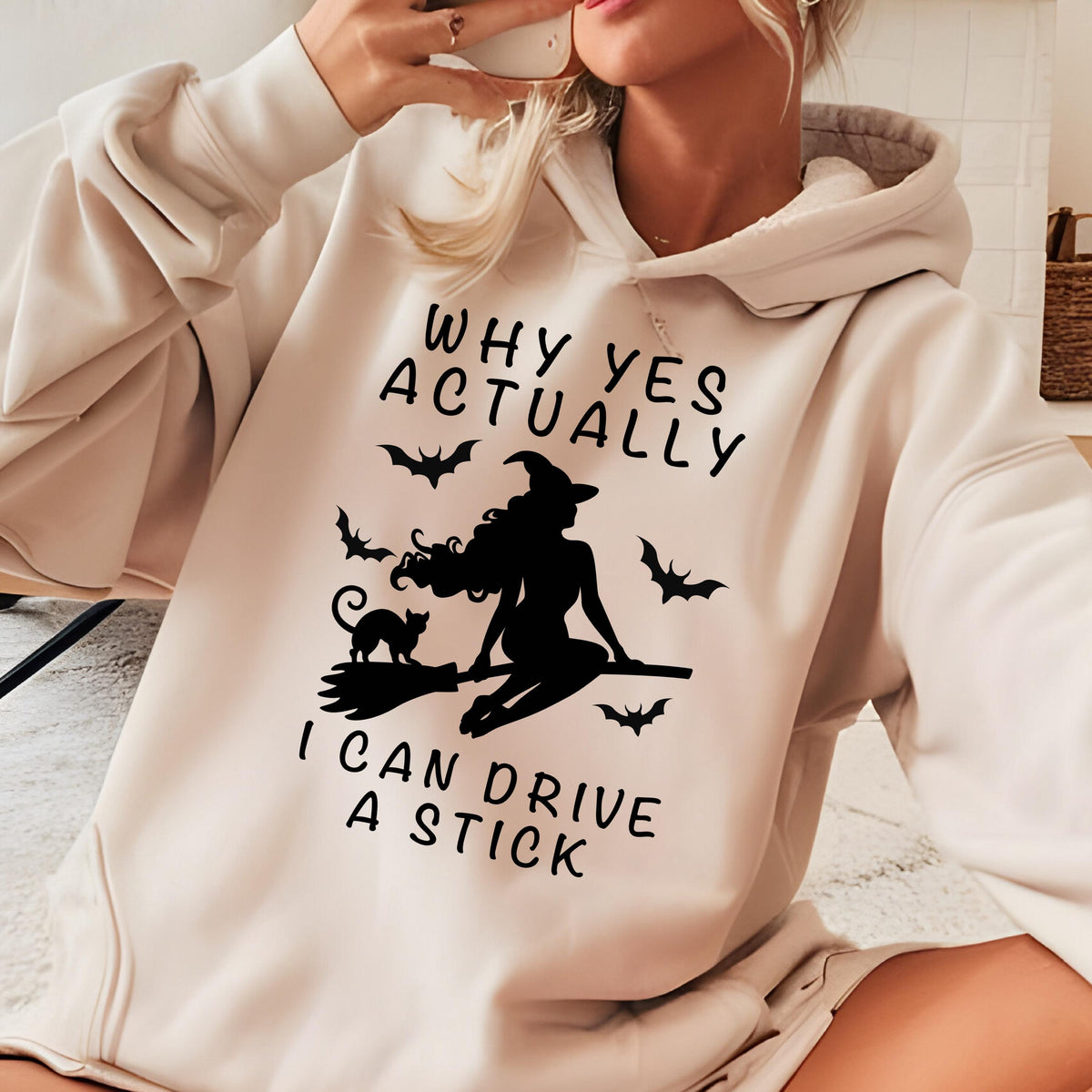 May include: A beige hooded sweatshirt featuring a black graphic print of a witch riding a broomstick with a cat and bats. The text reads: WHY YES ACTUALLY I CAN DRIVE A STICK. The garment has long sleeves, a front kangaroo pocket, and a drawstring hood.