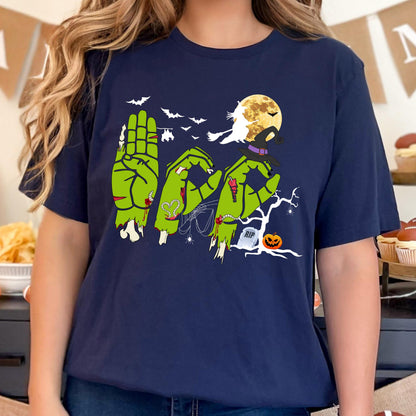May include: A navy blue t-shirt featuring a graphic of three green zombie hands forming the letters B, O, and O. The design includes a yellow full moon, a witch on a broomstick, bats, a spider web, a tombstone with RIP text, and a jack-o'-lantern.