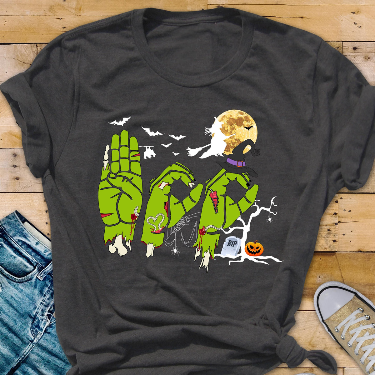 May include: A dark gray t-shirt featuring a graphic of three green zombie hands forming the letters B, O, and O. The design includes a yellow full moon, a silhouette of a witch on a broomstick, bats, a spider web, a tombstone with RIP, and a jack-o'-lantern.