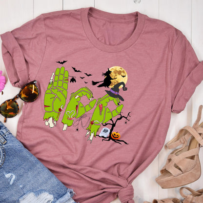 May include: A mauve short-sleeved t-shirt featuring a graphic of green zombie hands forming the word BOO. The design includes a witch on a broomstick, a full moon, bats, a tombstone with RIP text, a jack-o'-lantern, and a witch hat.