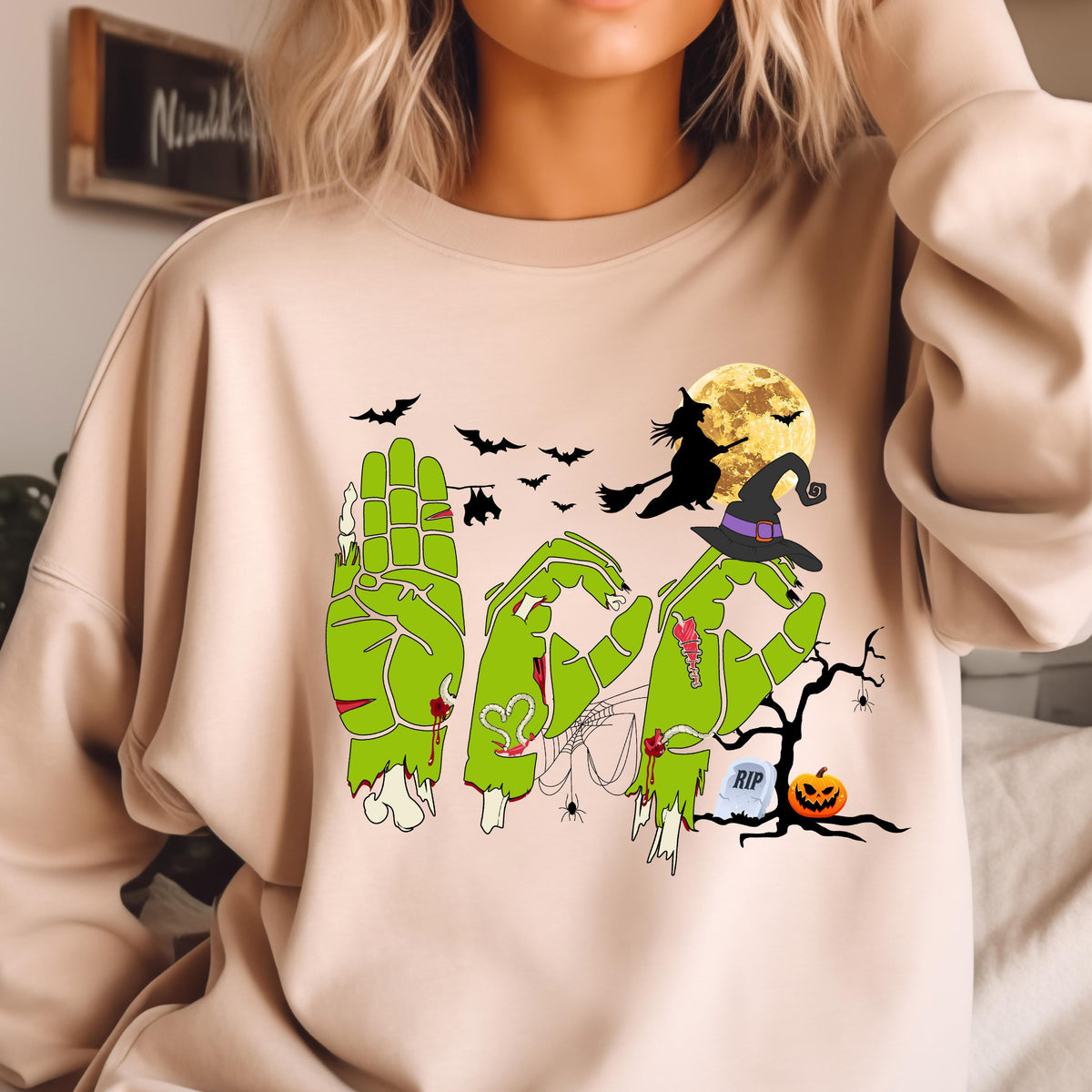 May include: Beige sweatshirt with a Halloween-themed graphic. The graphic spells out the word "BOO" using green zombie hands. A witch on a broom, bats, a full moon, a witch hat, a tree, a pumpkin, and a tombstone are also featured.