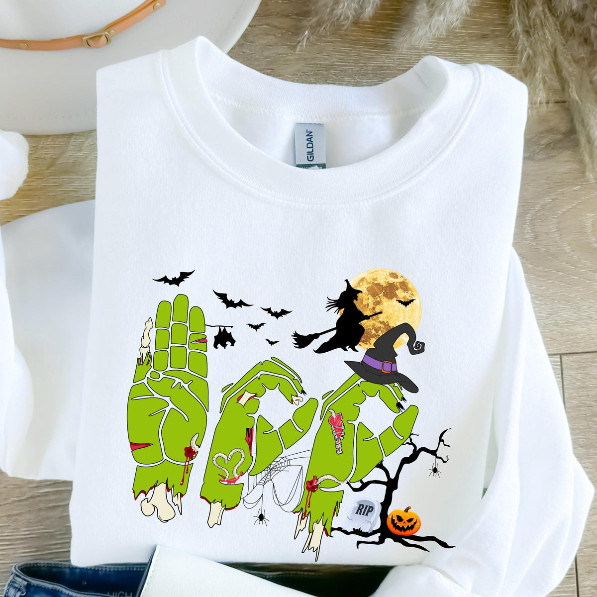 May include: White sweatshirt with a Halloween-themed graphic. The design features green zombie hands spelling out the word "BOO", a witch on a broom, bats, a full moon, and a jack-o'-lantern. The word "RIP" is also included in the design.