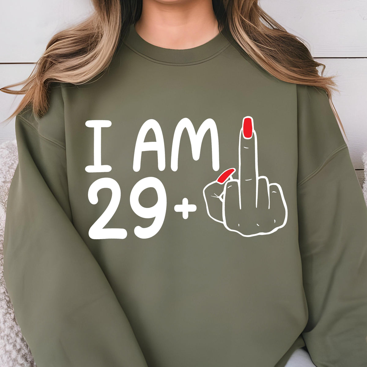 May include: Olive green sweatshirt with white text that reads "I AM 29+." A white outline of a hand giving the middle finger is to the right of the text. The middle finger has red nail polish.