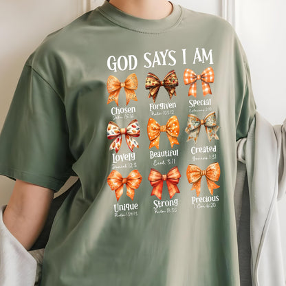 May include: An olive green t-shirt featuring the text "GOD SAYS I AM" above nine illustrated bows in shades of orange and brown. Each bow is paired with a descriptive word and a Bible verse reference, including Chosen, Forgiven, Special, Lovely, Beautiful, Created, Unique, Strong, and Precious.