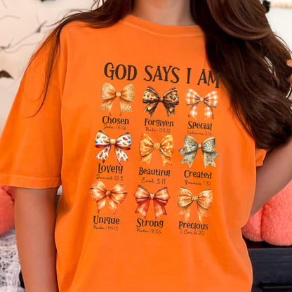 May include: An orange t-shirt featuring the text "GOD SAYS I AM" above nine distinct graphic bows. Each bow is paired with a descriptive word and a Bible verse reference, including "Chosen," "Forgiven," "Special," "Lovely," "Beautiful," "Created," "Unique," "Strong," and "Precious."