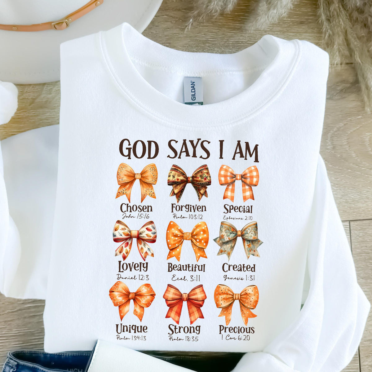 May include: A white crewneck sweatshirt featuring the text GOD SAYS I AM in brown letters. Below the text are nine illustrations of bows in various orange and brown patterns, each paired with a word and a Bible verse reference underneath.