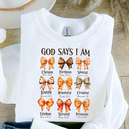 May include: A white crewneck sweatshirt featuring the text GOD SAYS I AM in brown letters. Below the text are nine illustrations of bows in various orange and brown patterns, each paired with a word and a Bible verse reference underneath.