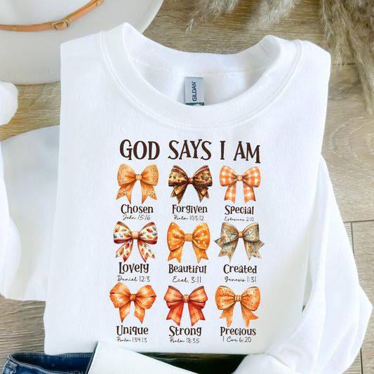 May include: A white crewneck sweatshirt featuring the text GOD SAYS I AM in brown letters. Below the text are nine illustrations of bows in various orange and brown patterns, each paired with a word and a Bible verse reference underneath.