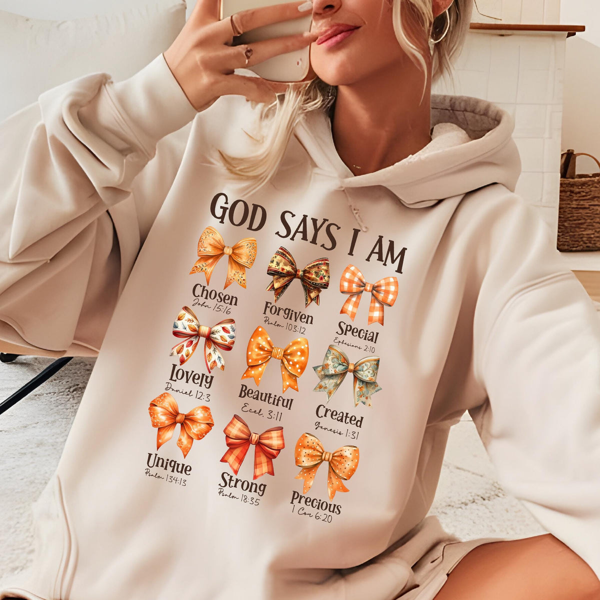 May include: A beige hooded sweatshirt featuring the text "GOD SAYS I AM" above nine colorful illustrated bows. Each bow is paired with a descriptive word and a Bible verse reference, including "Chosen," "Forgiven," "Special," "Lovely," "Beautiful," "Created," "Unique," "Strong," and "Precious."