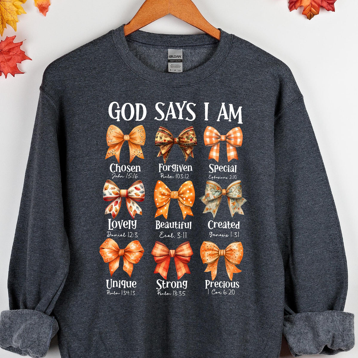 May include: A dark heather gray crewneck sweatshirt featuring the text GOD SAYS I AM at the top. Below are nine illustrated bows in various orange and brown patterns, each paired with a word and scripture reference like Chosen, Forgiven, and Special.