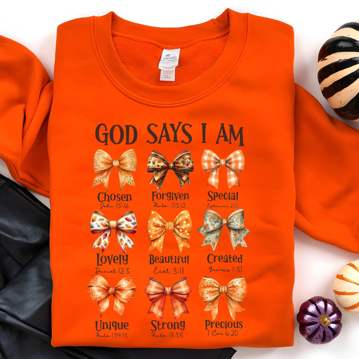 May include: A bright orange crewneck sweatshirt featuring the text "GOD SAYS I AM" above a grid of nine illustrated bows. Each bow is paired with a descriptive word and a Bible verse reference, including "Chosen," "Forgiven," "Special," "Lovely," "Beautiful," "Created," "Unique," "Strong," and "Precious."